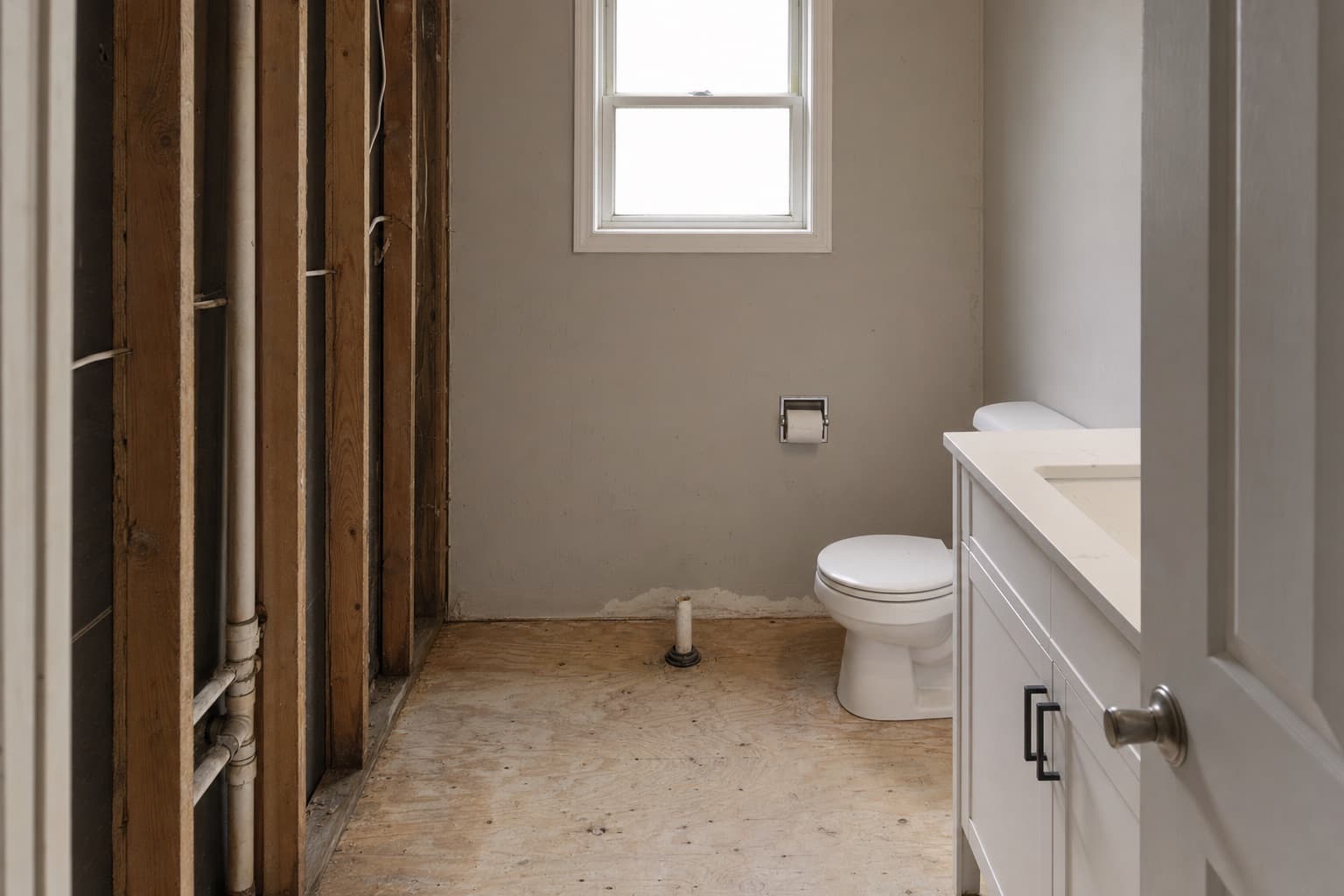 The same bathroom stripped to bare plywood subfloor with the drain stubbed up and studs exposed